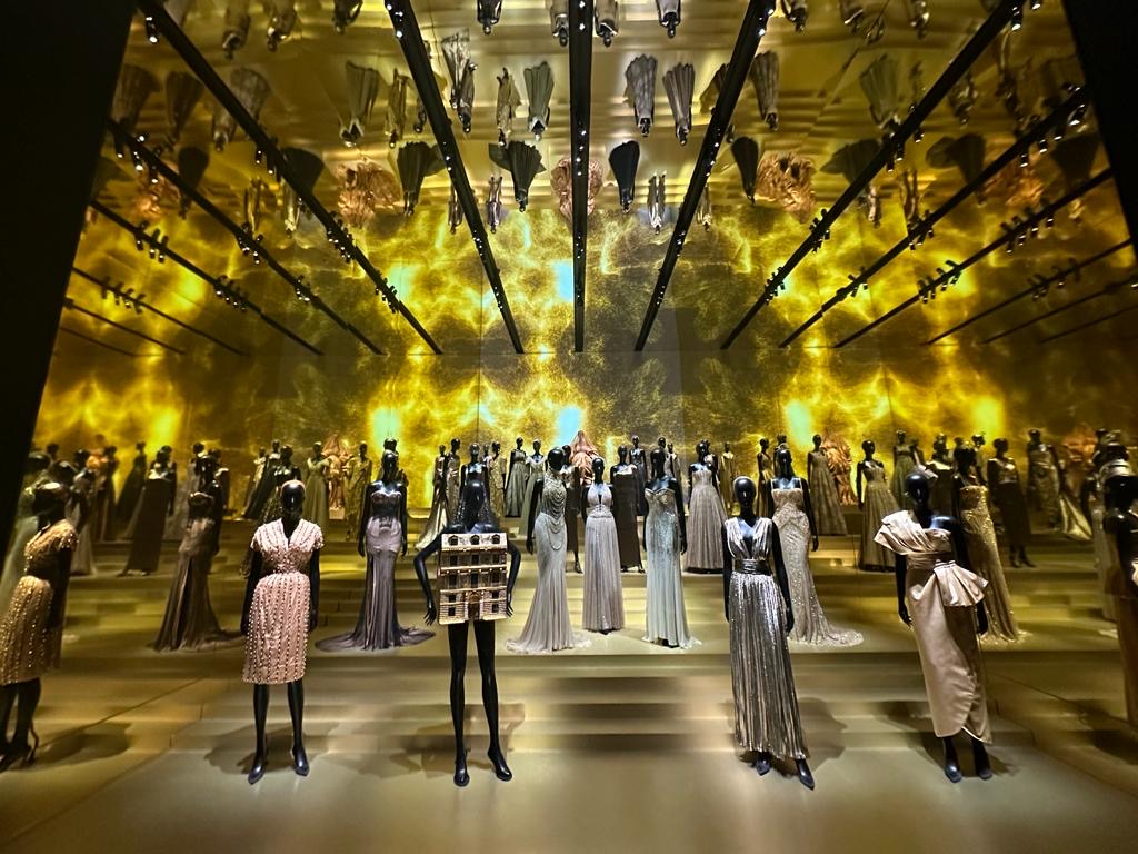 Univers totalement féérique en or dans une salle en verre qui permet d'avoir un reflet au plafond avec plein de faux mannequins qui pausent habillés avec des robes mythiques de Dior