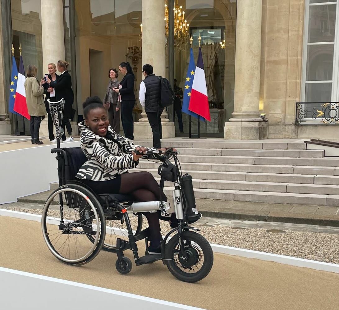 Deza femme noir, 2lègante dans son fauteuil roulant, pose devant le Palais présidentiel de l'Elysée lors d'une conférence nationale du handicap. Son expression dénote détermination et confiance, symbolisant l'inclusion et la représentation au cœur de cet évènement. 