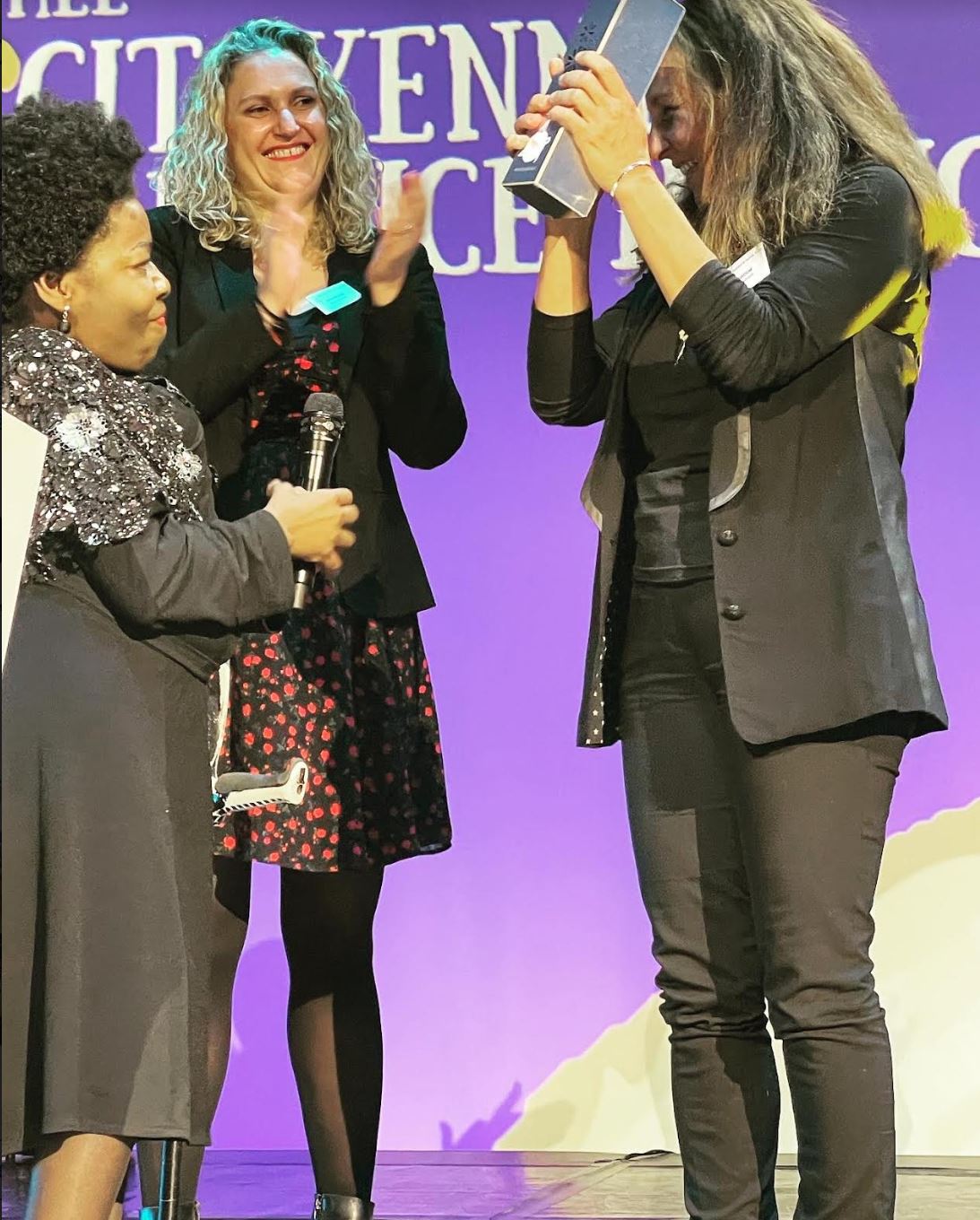 Deza, une femme de peau noire sourit largement alors qu'elle remet un trophée à une autre personne, capturant un moment de joie et de célébration. 