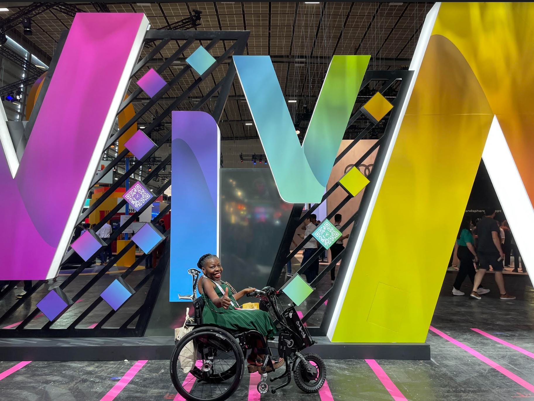 Une scène capturant une femme noire handicapée dans un fauteuil roulant à Viva teck, Face à une installation imposante portant le mot "VIVA" avec des QR codes, à l'accueil. La diversité et l'inclusion semblent être mises en avant dans cet évènement à la porte de Versailles 